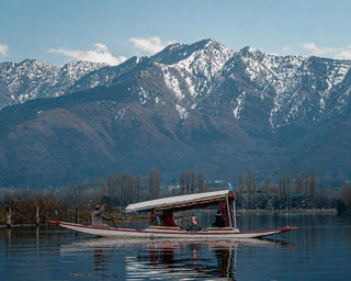 A traditional shikara boat in Kashmir with a canopy floats on calm blue water, with snow-capped mountains and a tree-lined shore in the background under a clear sky.